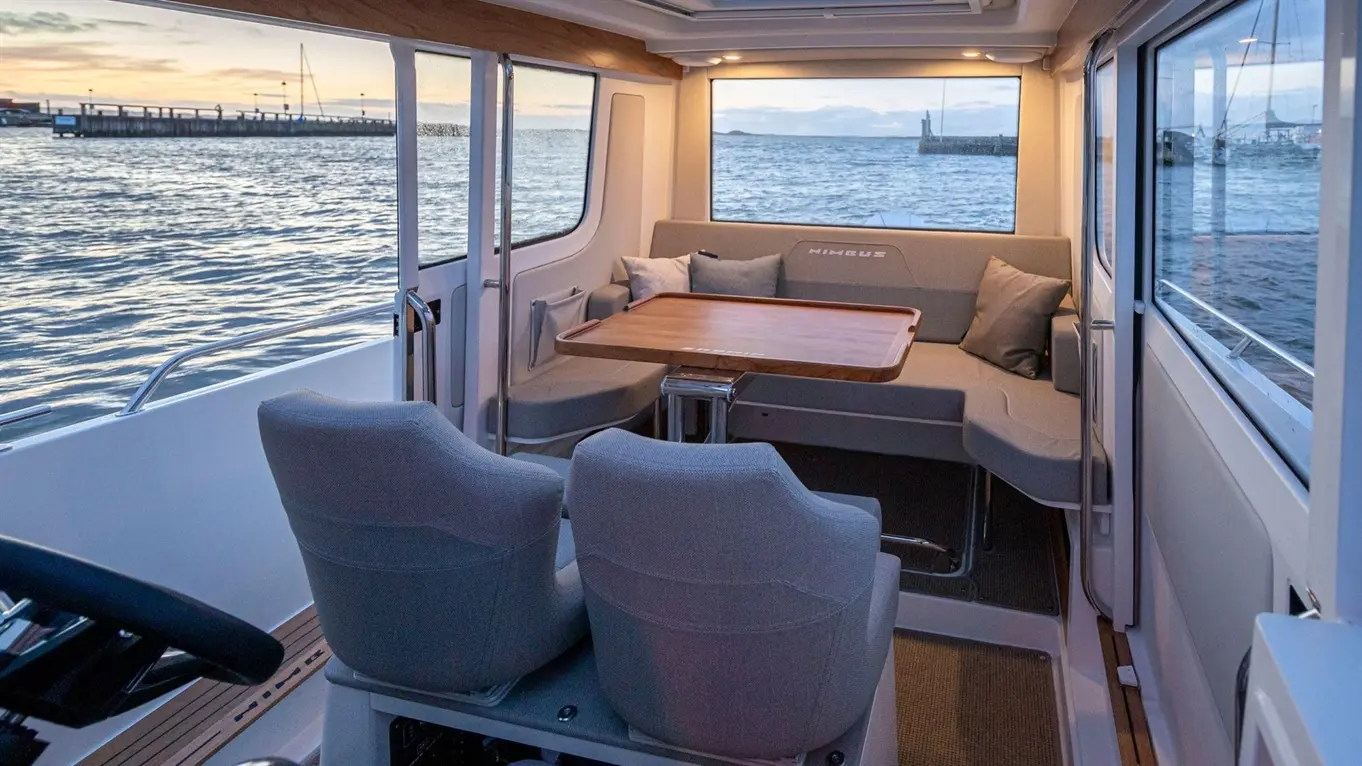 Interior of a modern boat featuring a dining table, two grey chairs, and views of the water and pier outside.